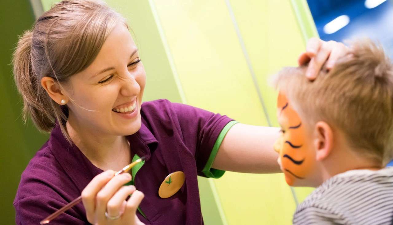 Face painter smiles while applying brush strokes to a child’s cheek, creating tiger stripes; indoors with bright green walls and soft lighting, child seated and facing away.