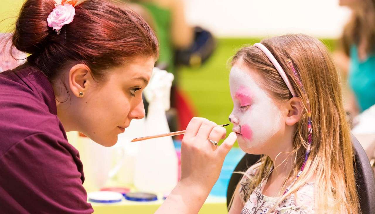 Face painter applies brush to a girl's cheek, finishing white-and-pink face paint. They concentrate closely across a table, with paint pots and blurred people in a bright indoor event.