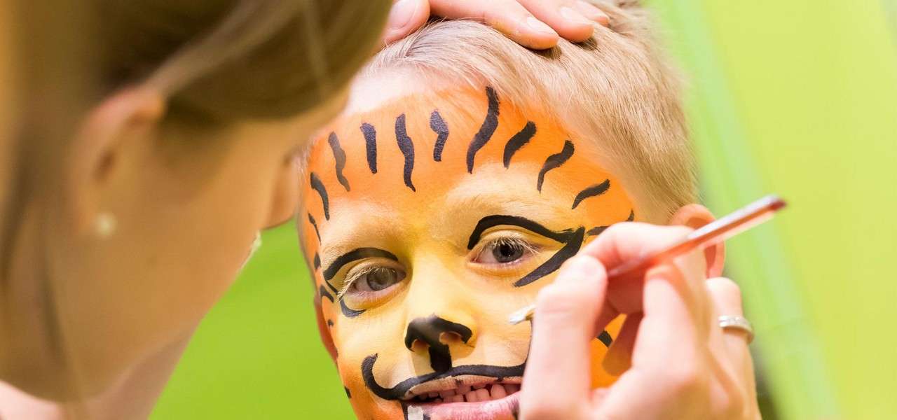 Child’s face—painted as a tiger—receives finishing touches; an artist holds the head and applies paint with a small brush against a bright green background.