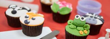 Animal-themed cupcakes display fondant toppers—a frog on green, fox-like face, panda, zebra, and flower—arranged on a red work surface beside decorating tools like cutters and a palette knife.