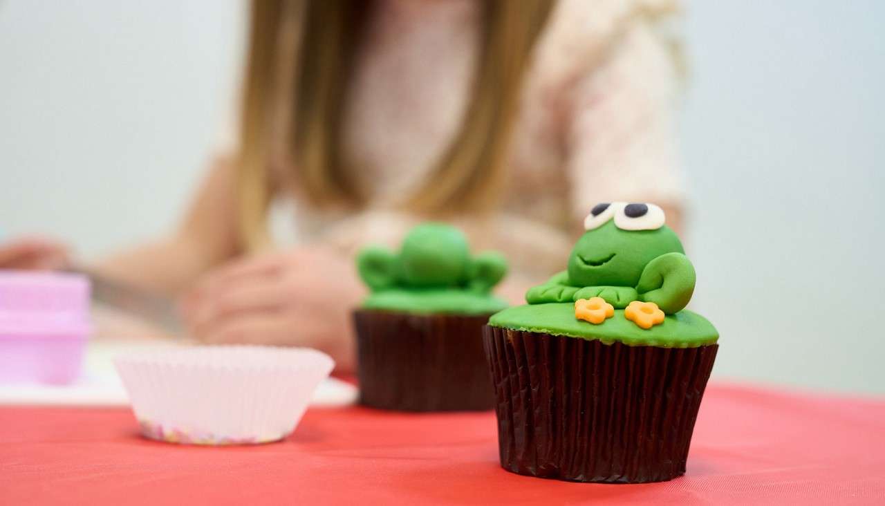Frog-topped cupcake sits on a red tablecloth, another decorated cupcake behind it. A child with long hair works in the blurred background near baking cups and tools.