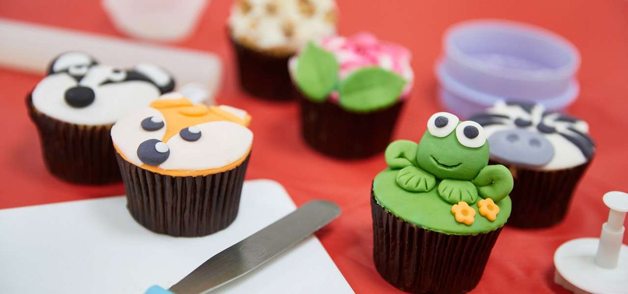 Cupcakes display animal-themed fondant decorations, including a frog, zebra, fox, and bear, sitting on a red work surface with a palette knife, cutters, and plastic molds scattered around.