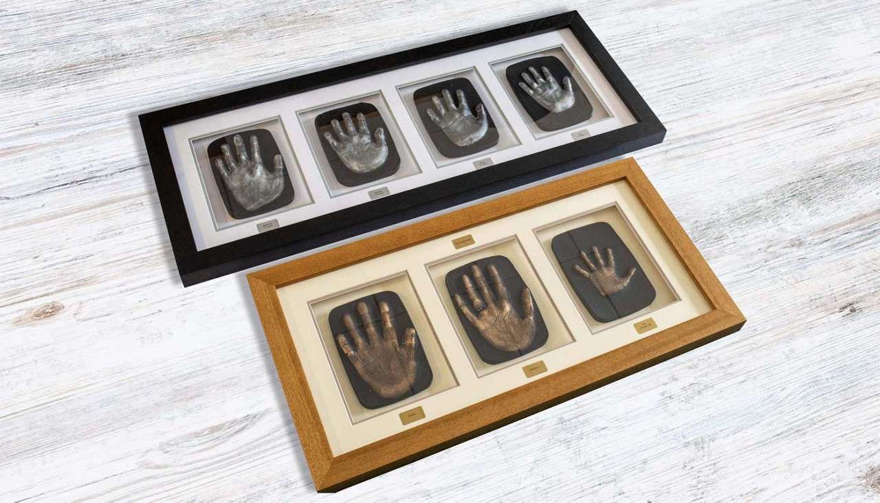 Framed hand-cast impressions rest on a white wooden surface. A black frame holds four silver palms; a wood frame holds three bronze palms, different sizes, each above small plaques with illegible text.