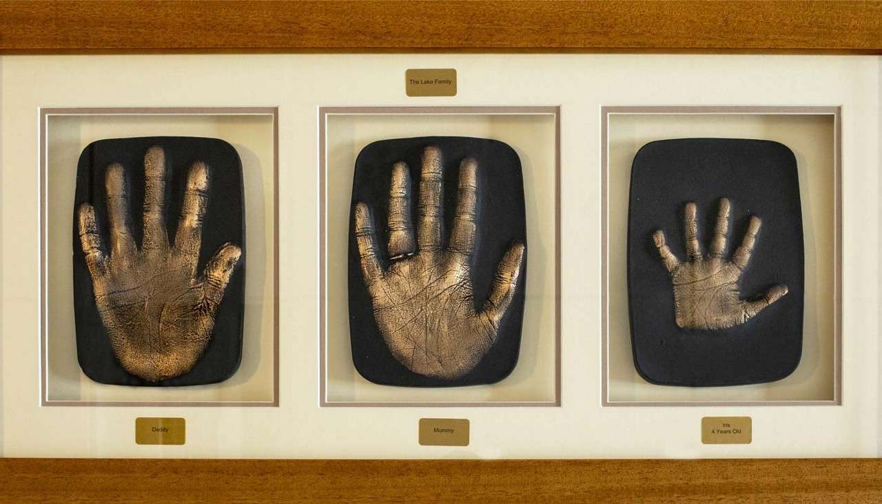 Three metallic handprint casts rest in a wooden frame, displayed side by side on a wall. Nameplates read: The Lake Family; Daddy; Mummy; Iris 4 Years Old.