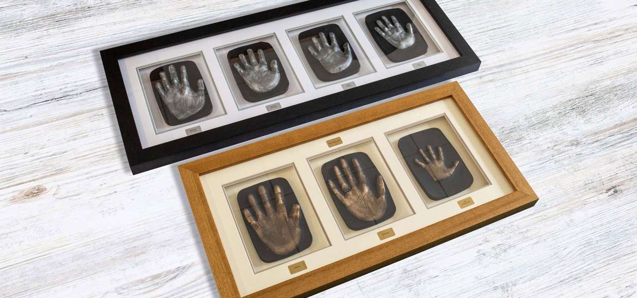 Framed handprint casts—metallic impressions—sit aligned in two rectangular frames. The upper black frame holds five prints; the lower wooden frame holds three. Both lie on a whitewashed wooden surface.