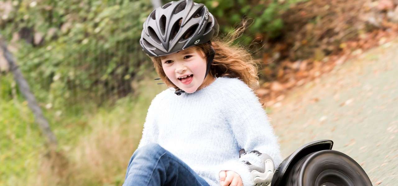 Child in a bike helmet rides a low wheeled go-kart, leaning and smiling; motion blur on front wheel. Background shows a paved path bordered by greenery, fence, and scattered leaves.