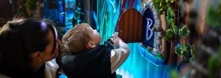 Child reaches into a small arched door to reveal a round chalkboard marked “B”, as an adult watches beside, within a dim, colorful, plant-decorated interactive exhibit.