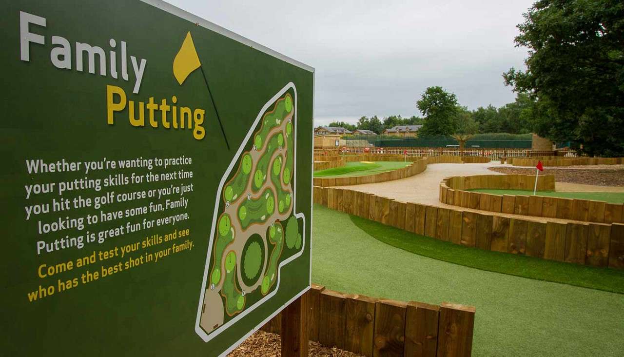 Signboard advertises family putting, standing beside a landscaped mini‑golf course with wooden borders and artificial turf. Text: “Family Putting. Whether you’re wanting to practice your putting skills for the next time you hit the golf course or you’re just looking to have some fun, Family Putting is great fun for everyone. Come and test your skills and see who has the best shot in your family.”