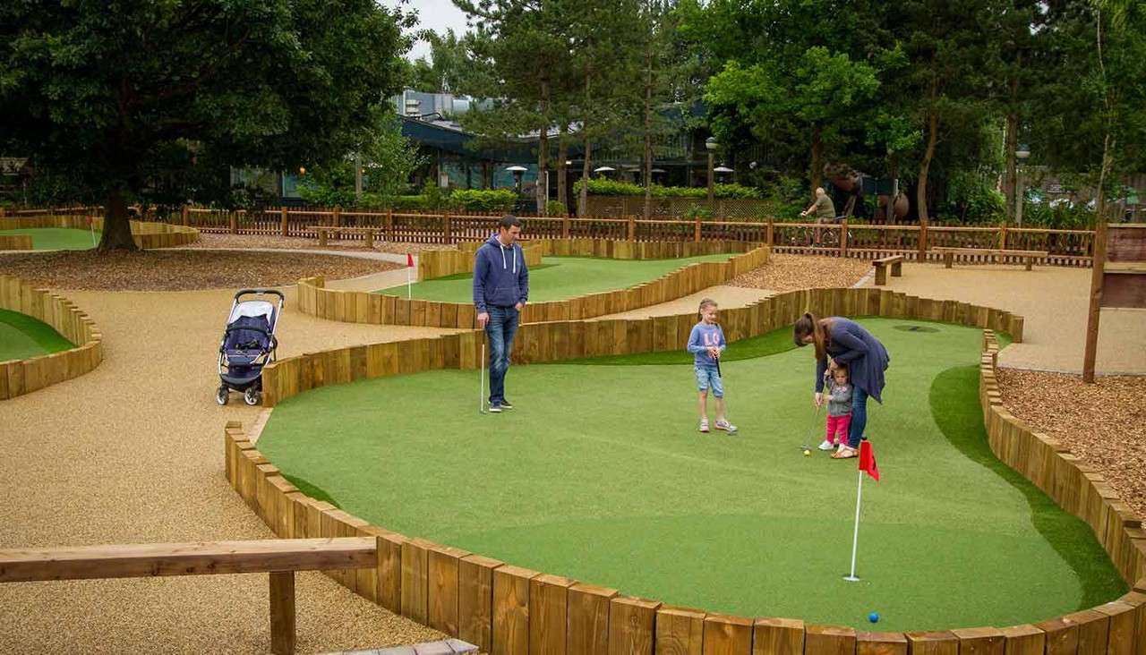 Family plays miniature golf; an adult guides a toddler’s putt while another adult and child watch. Context: artificial-turf greens with wooden edging, red flags, gravel paths, stroller, trees, and fences.