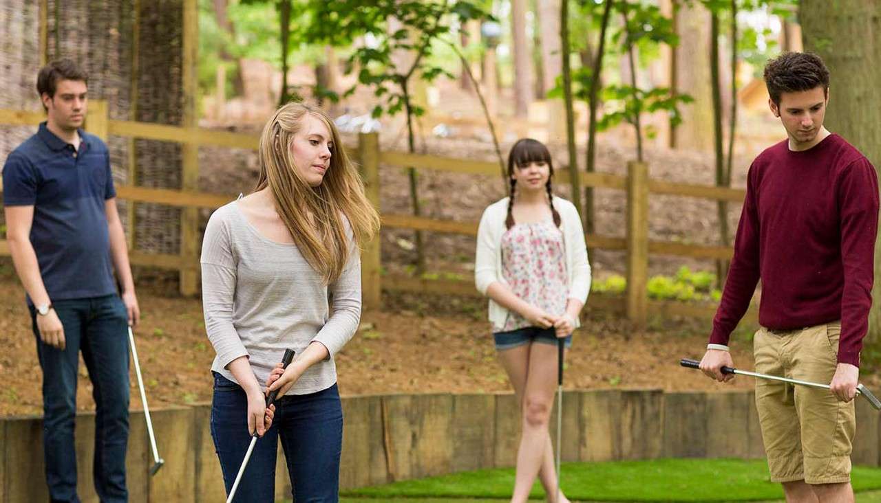 Four young adults with putters pause and look around on a mini-golf green; two stand foreground, two background; wooded park setting with trees, mulch, and a wooden fence.