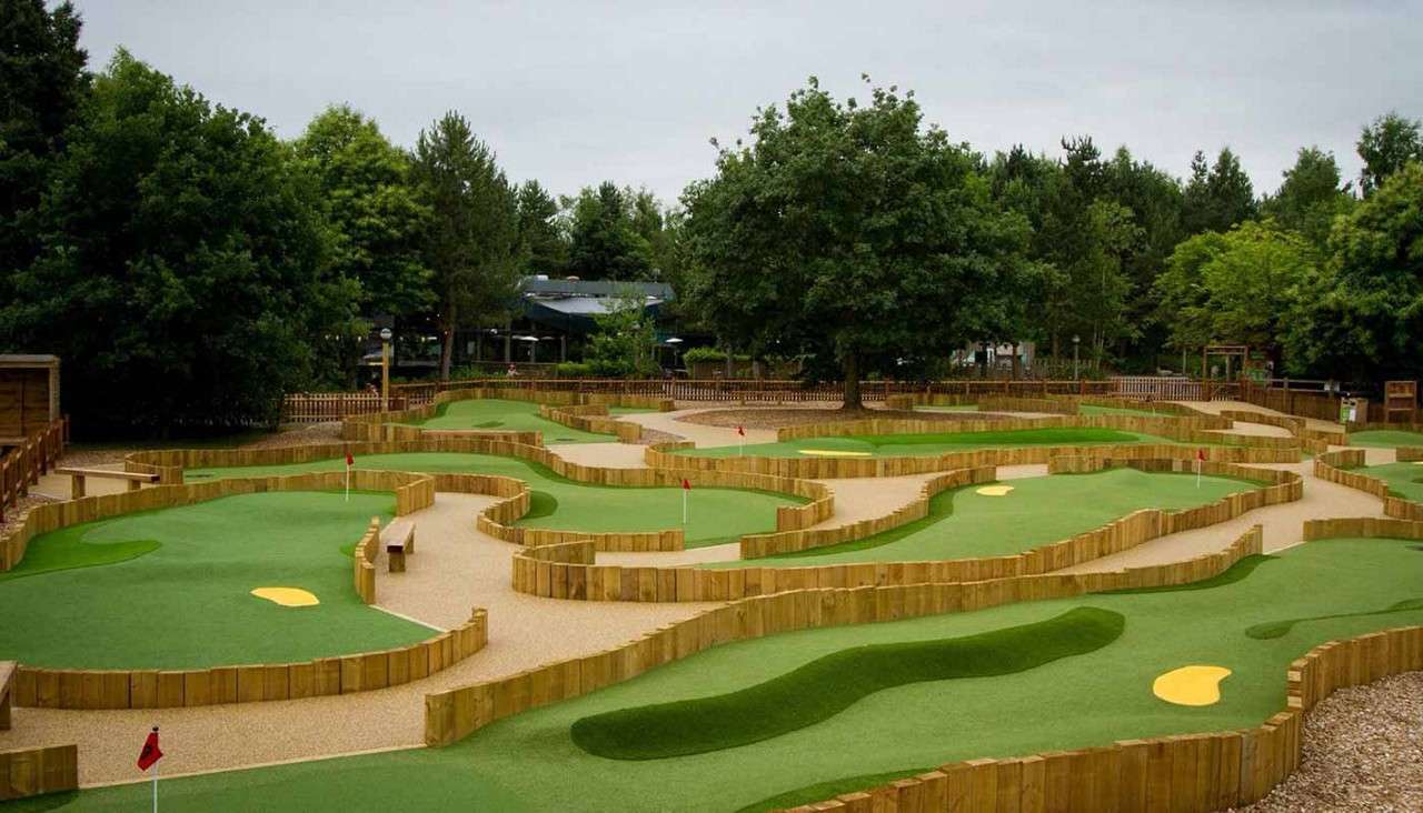 Mini-golf course features winding artificial greens with small red flags, bounded by low wooden walls, in a landscaped park setting surrounded by dense trees on a cloudy day.
