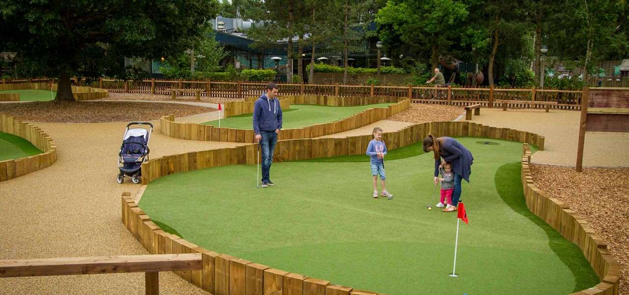 Family plays mini-golf; a woman helps a small child putt while a man and older child watch. Surrounding landscaped course features wooden borders, flag pins, stroller, trees, and fences.