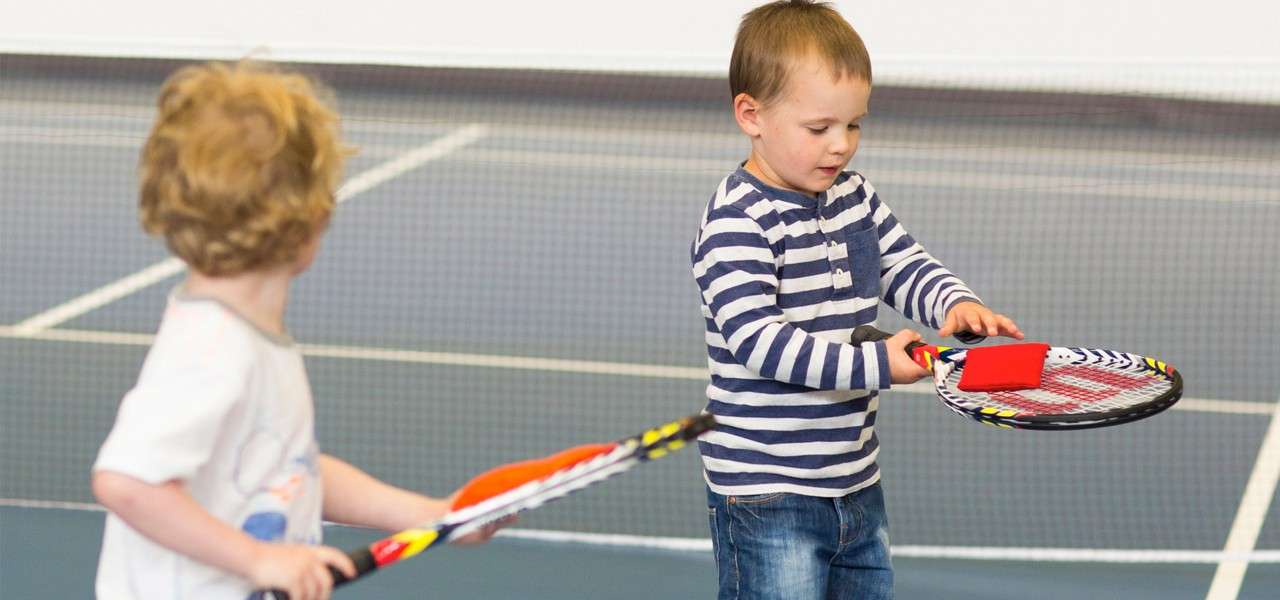 Two young children hold tennis rackets, balancing small red and orange beanbags on the strings, while practicing together on an indoor tennis court with a net and painted boundary lines.