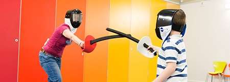 Two people in fencing masks duel with foam swords, one lunging while the other blocks. They stand indoors before a red-to-yellow paneled wall; a small orange chair sits nearby.