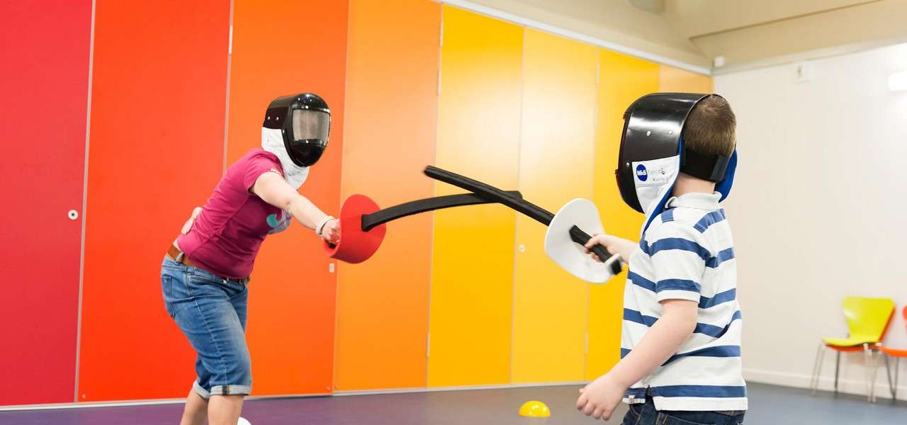 Two fencers spar with foam swords, wearing helmets and small shields, in a bright indoor room with orange-yellow wall panels, purple floor, and a few chairs in the background.