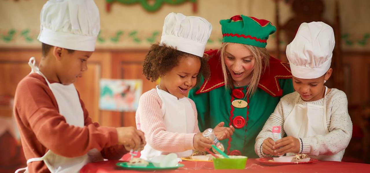 Children in chef hats decorate cookies with icing, guided by a smiling person in an elf costume, at a red-topped table in a warmly lit, Christmas-themed room.