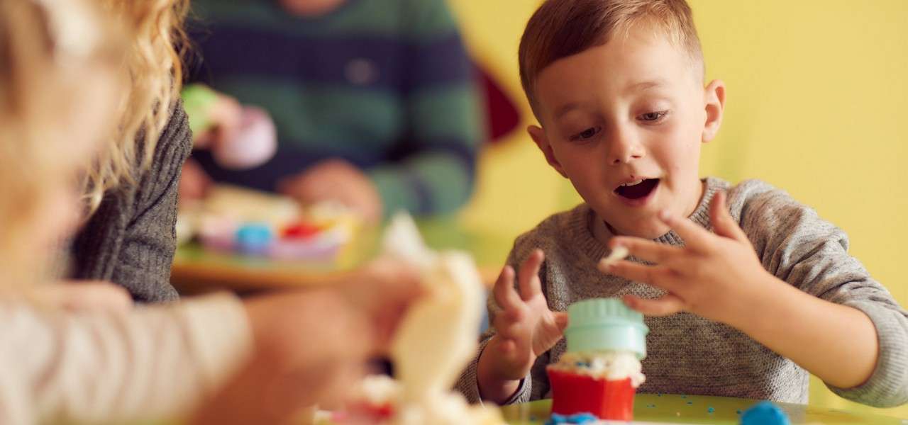Child decorates a cupcake, smearing frosting on fingers while positioning a blue silicone cup on top; context: colorful classroom party table with other children and crafting materials in soft focus.