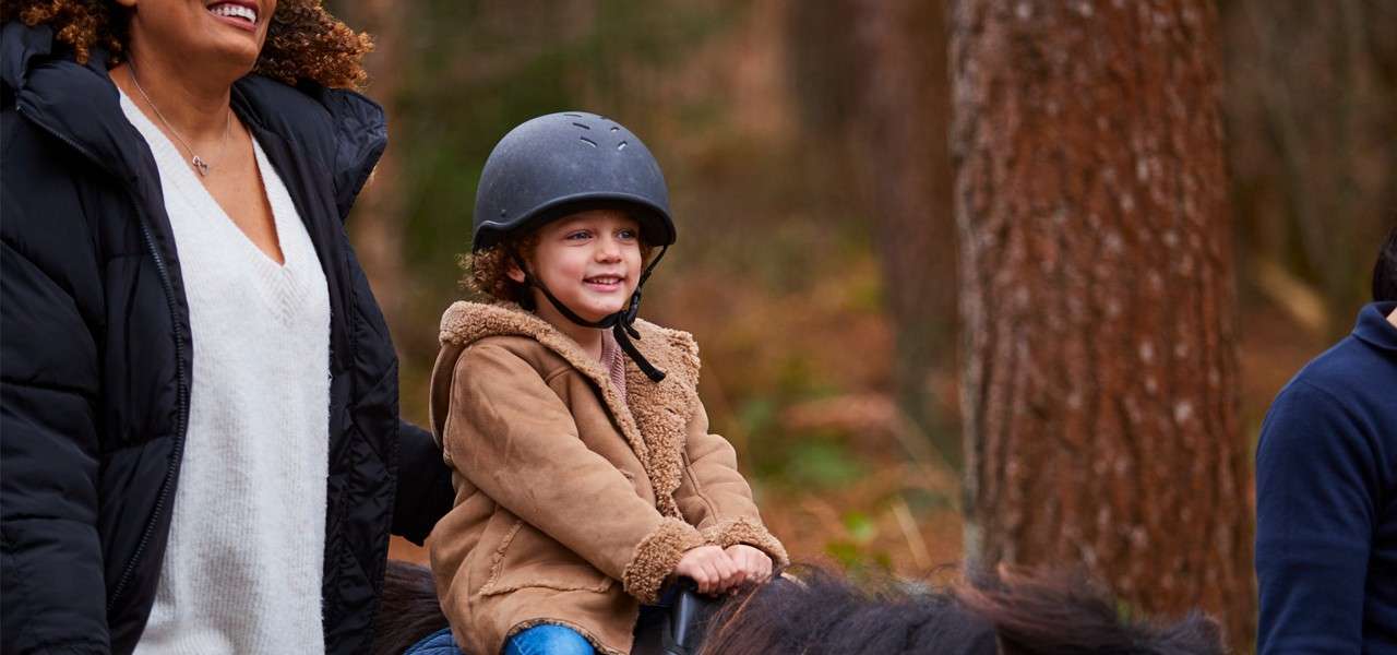 Child wearing a riding helmet smiles while holding reins, riding a pony; an adult walks beside them as they move along a forest trail lined with tall trees and a soft, earthy background.