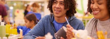 Two people paint ceramic mugs with thin brushes, concentrating at a shared table; paint bottles and tools surround them, with other participants and shelves blurred in a bright craft studio.