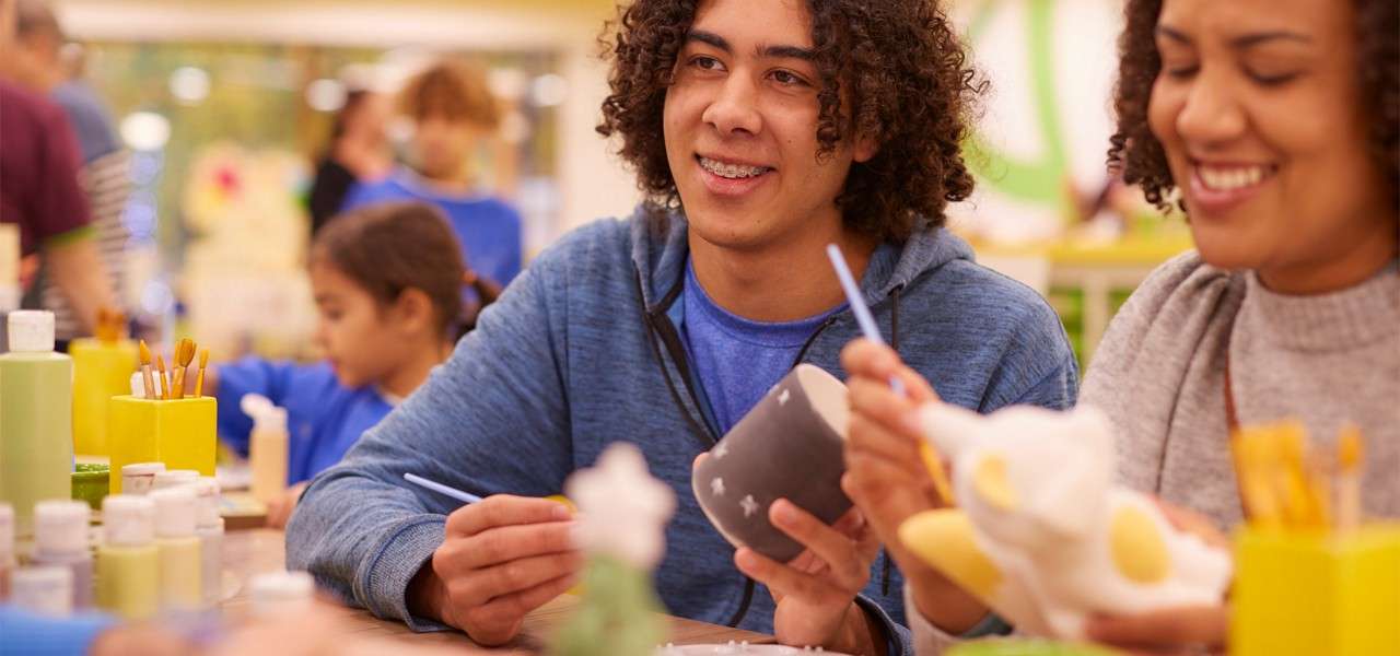 Smiling teen paints a gray ceramic mug with a brush, at a busy arts-and-crafts studio; others at the table paint ceramics, with paint bottles and brush holders scattered around.