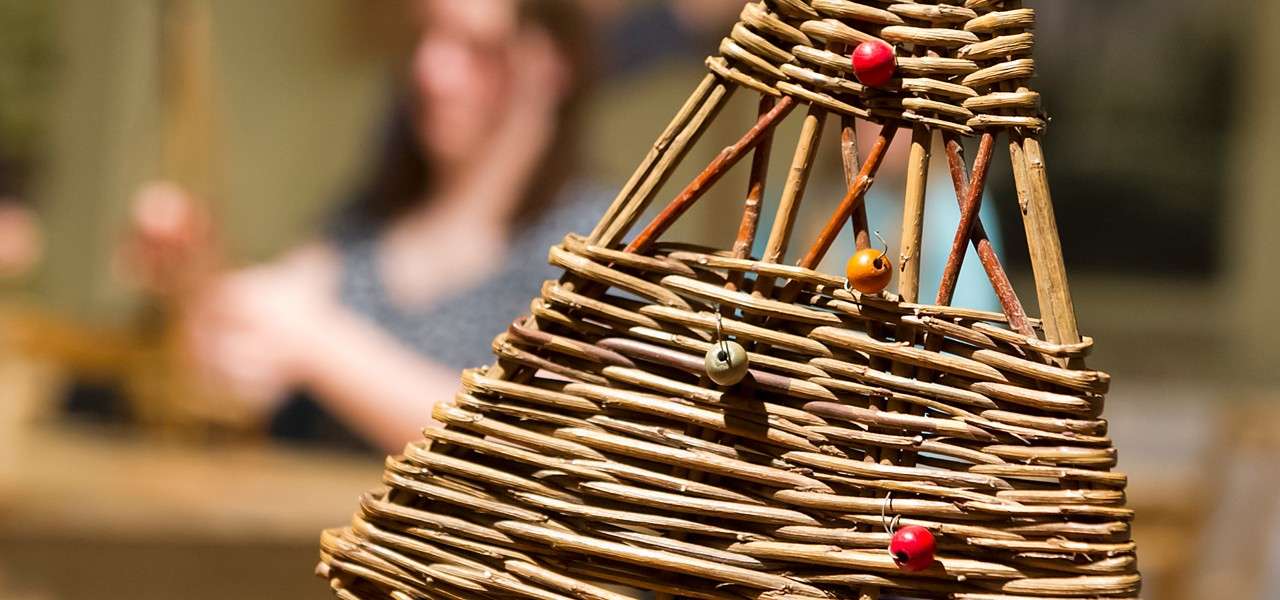 Wicker cone sculpture, decorated with small red, orange, and green beads, stands in focus; behind it, a blurred person works with reeds in a craft workshop setting.