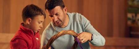 Man and boy handle a thick rope; the man demonstrates knot-tying while the boy watches closely, set inside a warmly lit wooden room.