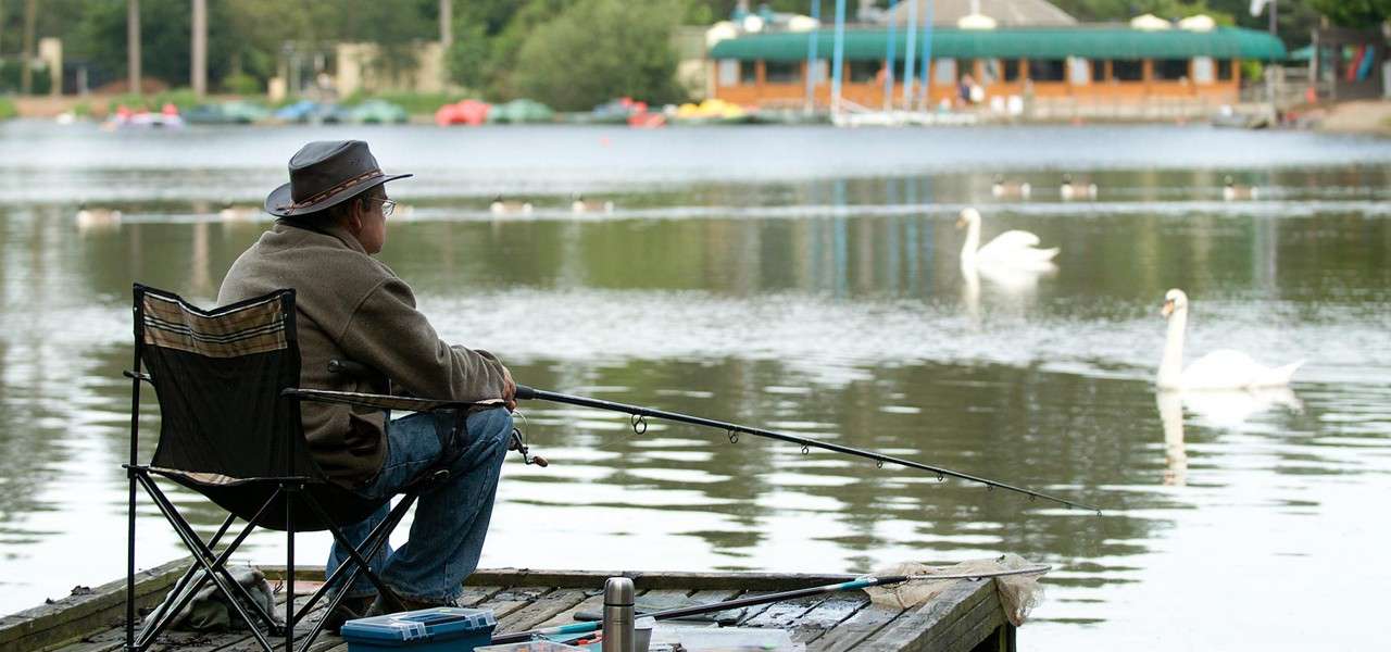 Man wearing hat sits on folding chair, holding a fishing rod, awaiting a bite, on a wooden dock by a calm lake with swans, moored boats, and lakeside buildings.