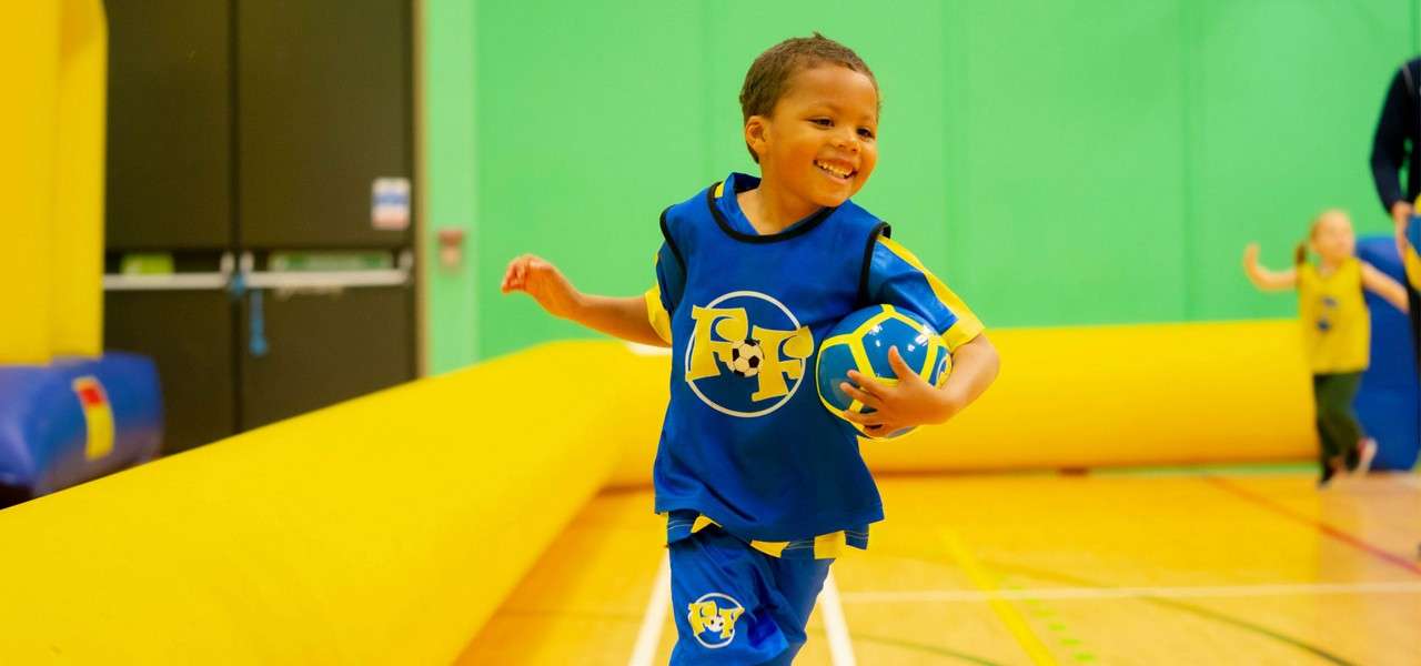 Child runs, cradling a blue-and-yellow ball, smiling. Wearing a blue sports kit with a circular logo. Indoors on a gym court bordered by yellow inflatables; other children play behind.