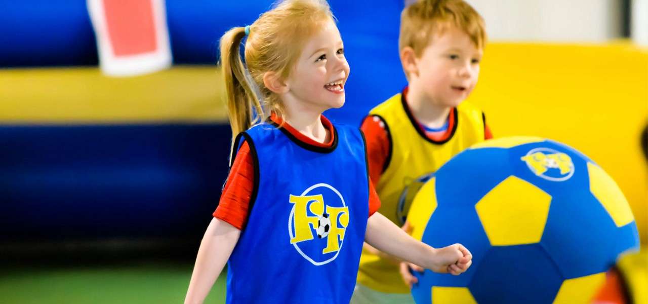 Children run and smile; one pushes a giant blue-yellow soccer ball. Indoor play area with colorful mats. Text: FF on the girl’s blue pinnie and repeated on the ball.