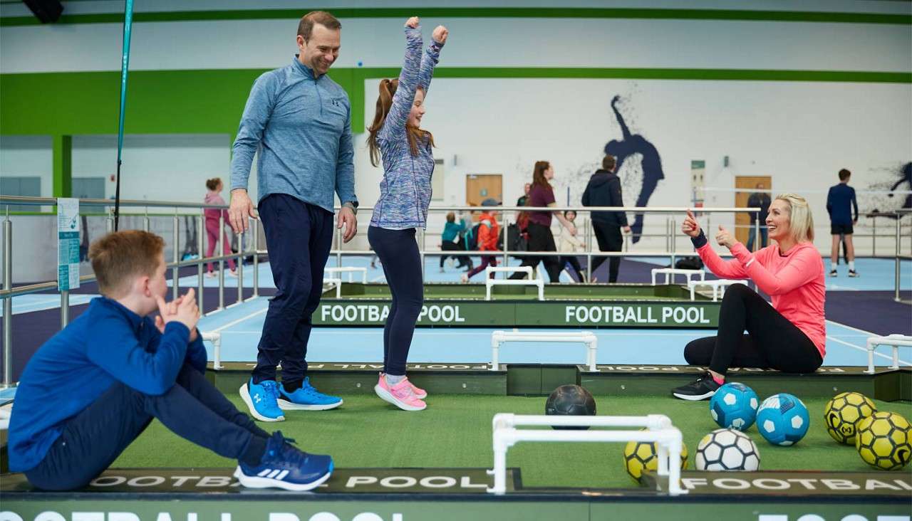 Family plays football pool; a girl celebrates with raised arms as adults encourage and a boy sits nearby; soccer balls dot a giant table in an indoor sports center. Text: FOOTBALL POOL.