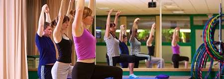 Group of adults raise arms while holding lunging poses, following a class leader. They stand on mats in a mirrored fitness studio with green walls and stacked colorful hula hoops.