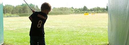 Child golfer swings a club; taking a shot from a covered bay toward an open driving range, grass and distance markers ahead. Jersey shows number 8; name above is unreadable.
