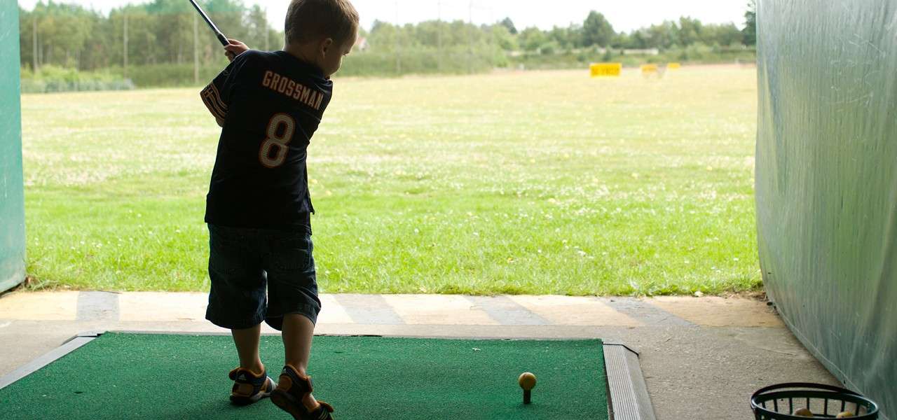 Child golfer swings a club at a yellow-teed ball on a practice mat, facing an open driving range with grass and distant target signs. Jersey text: GROSSMAN 8.