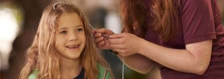Smiling child sits while an adult threads a beaded strand into their hair, hands near the head, in a softly blurred outdoor setting with trees.