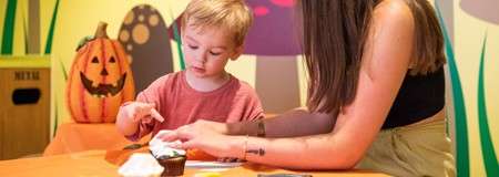 Child and adult craft with paper and glue, seated at a table; in a colorful, Halloween-themed playroom with a jack-o’-lantern, mushroom murals, and a trash bin labeled: WASTE.