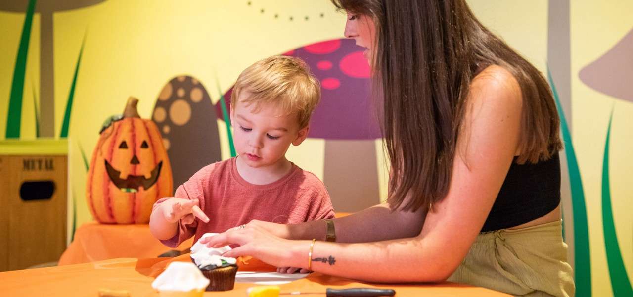 Child decorates a cupcake while an adult assists, both seated at an orange craft table; pumpkin jack-o’-lantern and cartoon mushroom mural create a cheerful indoor setting. MUTUAL.