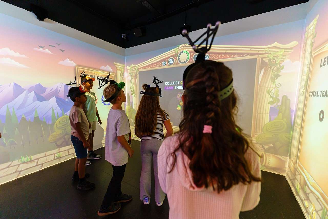Children wear headsets and watch an interactive wall projection in a gaming room with cartoon mountains; on-screen text: COLLECT, BANK THEM, TOTAL TEAM, 2x.