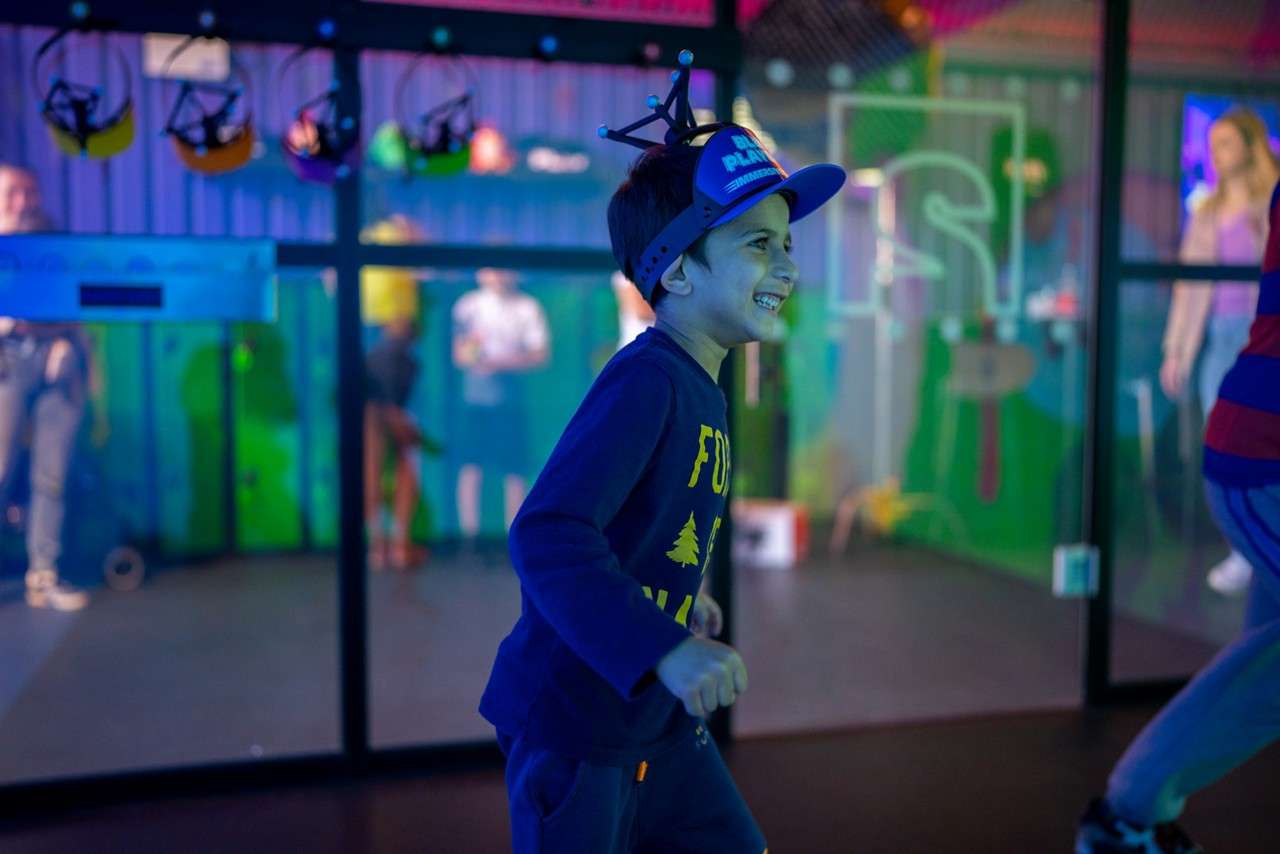 Child runs and grins, wearing a cap with tracking markers. In a neon-lit indoor play zone, people stand behind glass and gear hangs overhead. Visible text: PLAY; FO; S.
