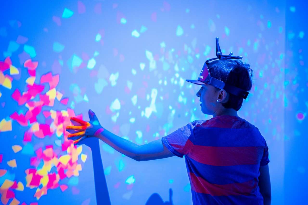 A child wearing a motion-tracker visor reaches toward a wall, triggering colorful projected confetti shapes; the blue-lit room surrounds them, casting a long hand shadow on the surface.