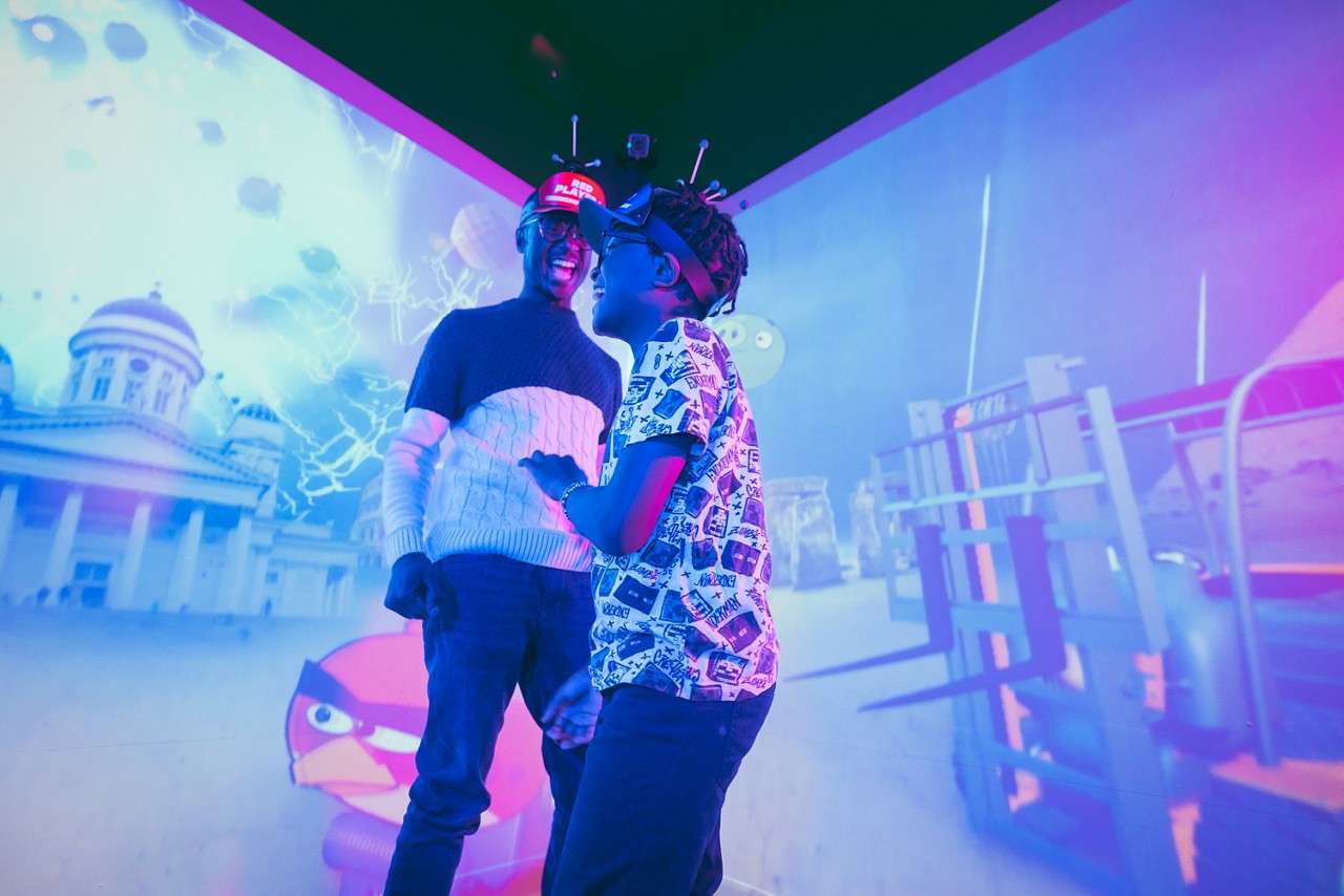 Two people in VR headsets laugh and interact, standing before a large, colorful animated projection with cartoon characters and city scenes. Visible text: RED PLAYER.