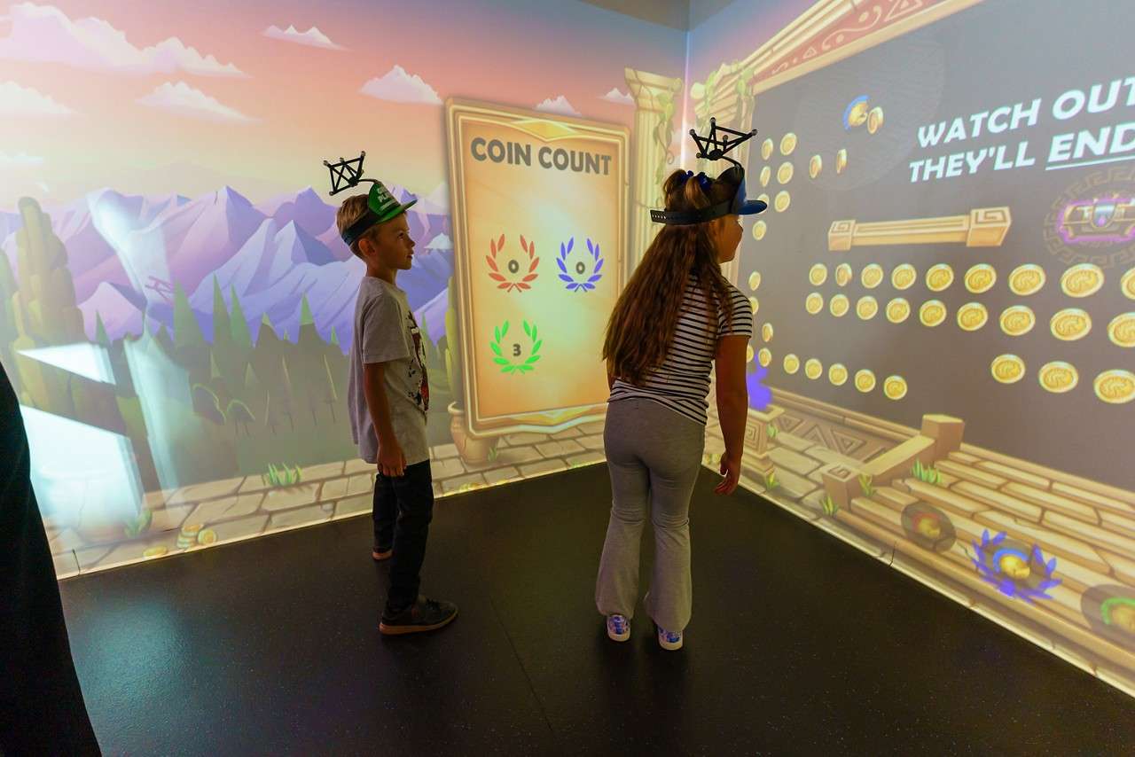 Two children wearing head-mounted trackers interact with a wall-projected coin-collecting game, moving in a small immersive room with cartoon mountains and a scoreboard.

Visible text: COIN COUNT; 0; 0; 3; WATCH OUT; THEY’LL; END…