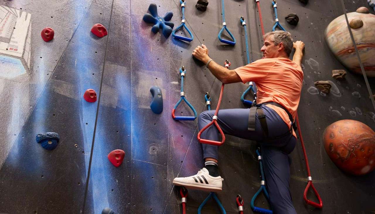 Climber ascends indoor rock wall, gripping rope loops and handholds; he wears harness, orange shirt, blue pants, white sneakers; space-themed mural with planets and a spaceship surrounds.