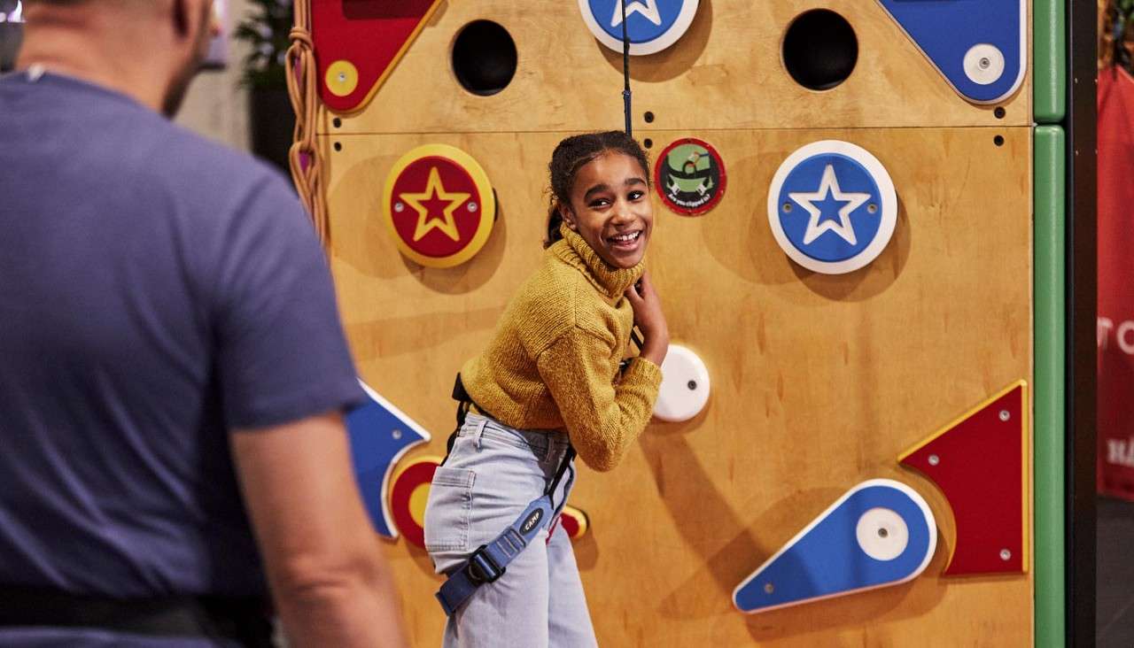 Girl climber smiles while wearing a harness, turning toward a supervising adult, attached to a top-rope on a colorful indoor climbing wall with circular holds and star-shaped panels.