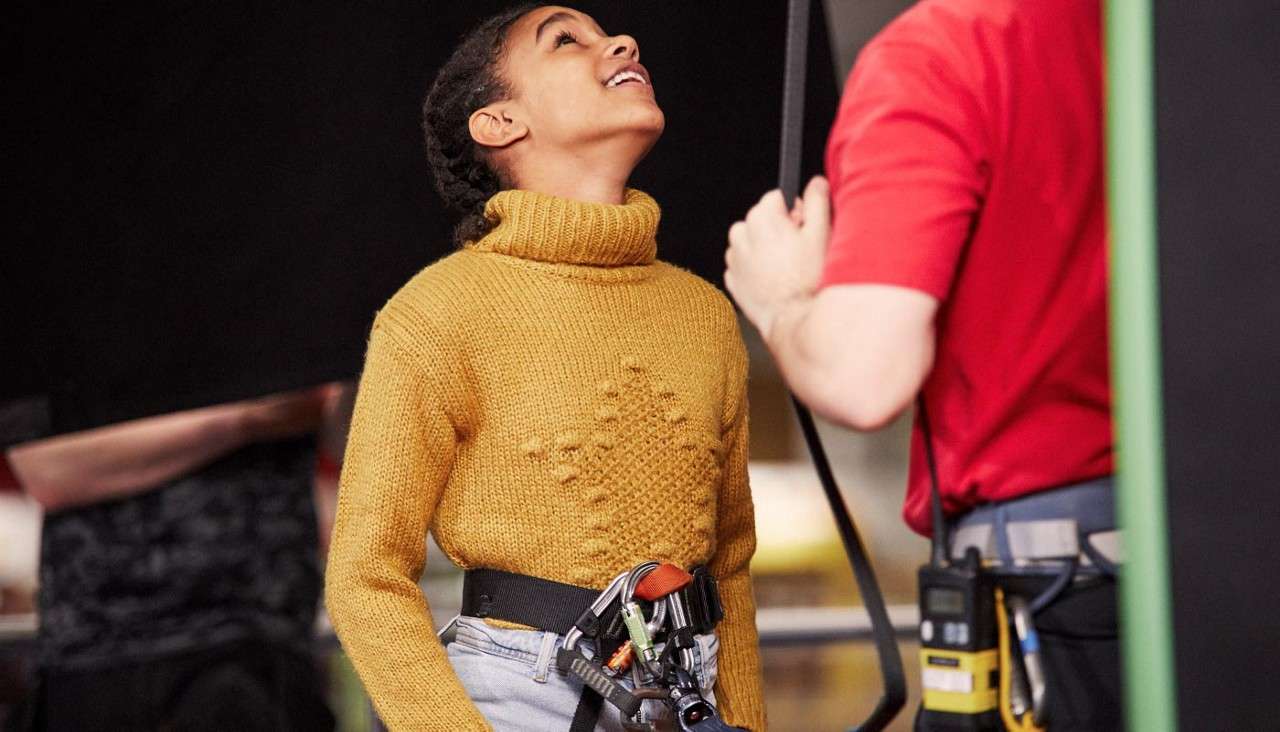 Child in a mustard turtleneck, wearing a climbing harness, looks upward while an instructor in a red shirt checks gear; indoor climbing or safety training environment.