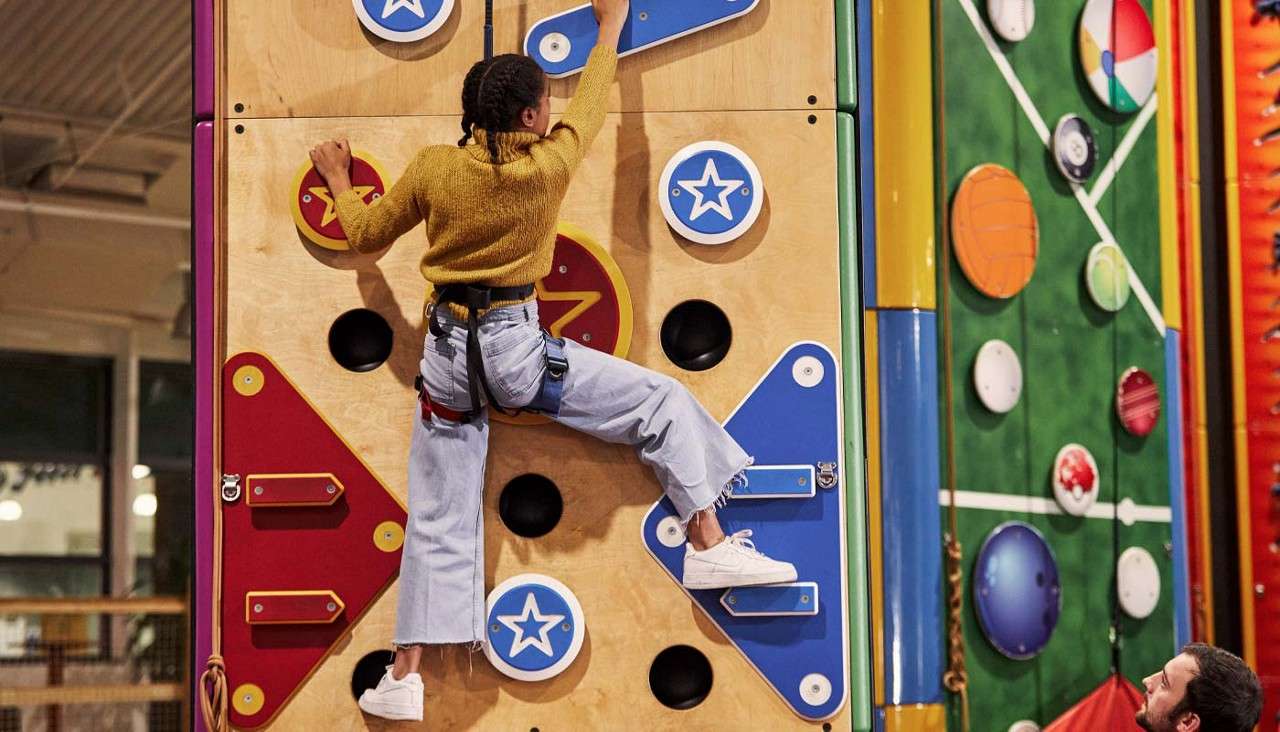 Child climber ascends a colorful indoor climbing wall, reaching for a blue hold. Harnessed, wearing jeans and sneakers. An adult watches below amid adjacent sports-themed walls in a recreation center.