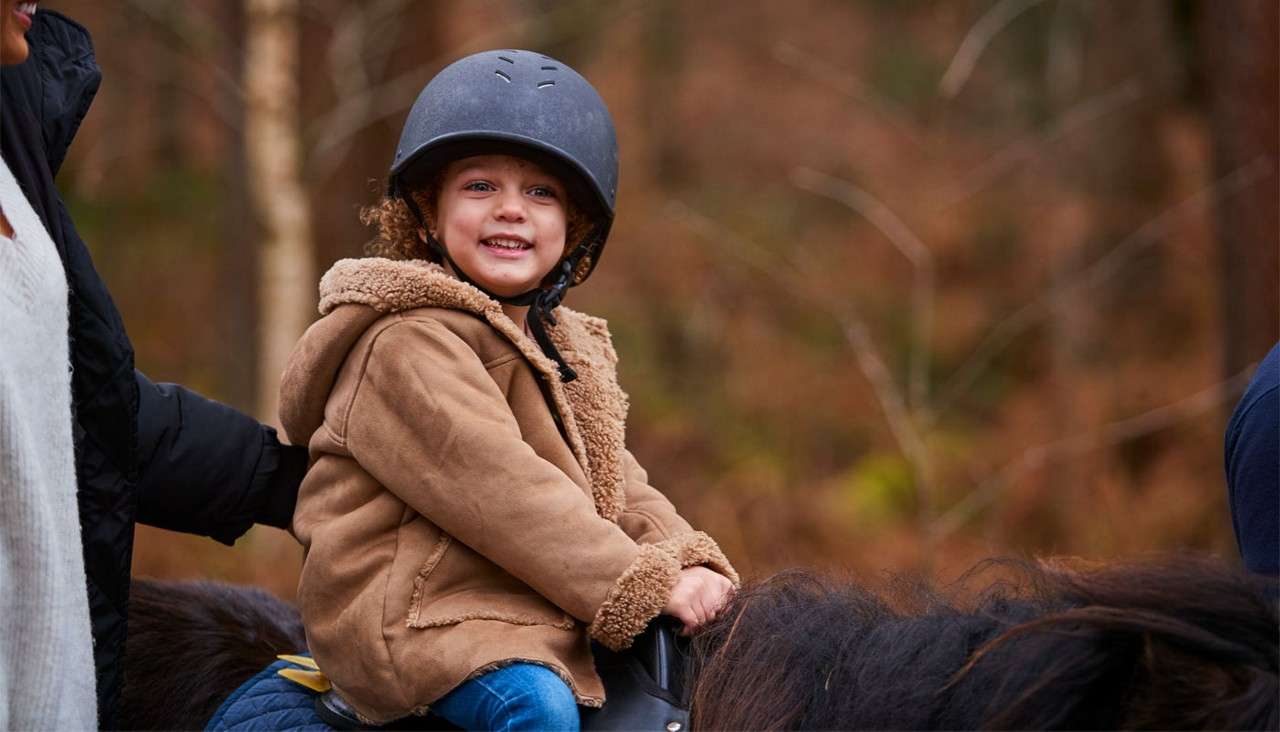 Child wearing a riding helmet smiles while sitting on a pony, holding the saddle; an adult steadies them. Background shows a blurred autumn forest, suggesting a guided trail ride.