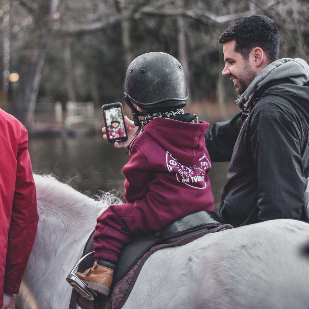 Child rider takes a selfie while seated on a white horse; an adult smiles beside them in a wooded park. Visible hoodie text: "Team" and "ON TOUR".
