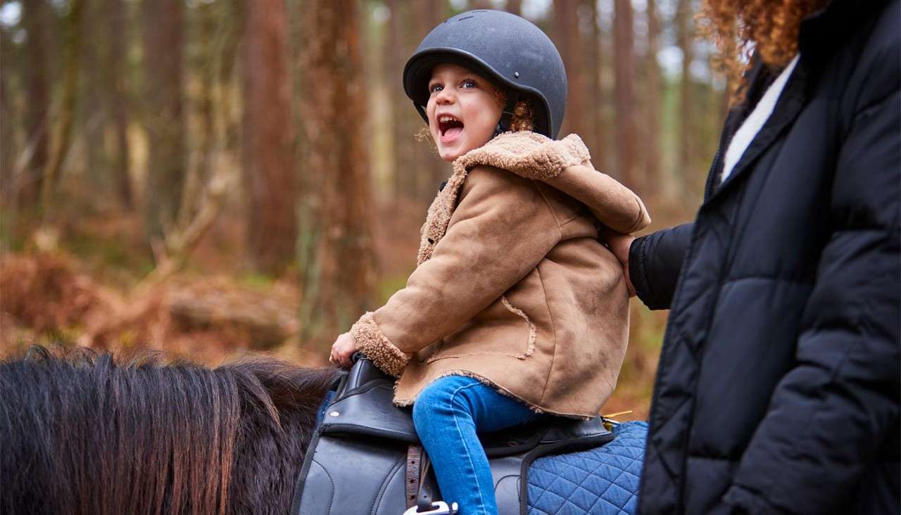 Child wearing a helmet rides a dark horse, smiling and looking back, while an adult steadies them at the saddle, within a wooded forest trail.