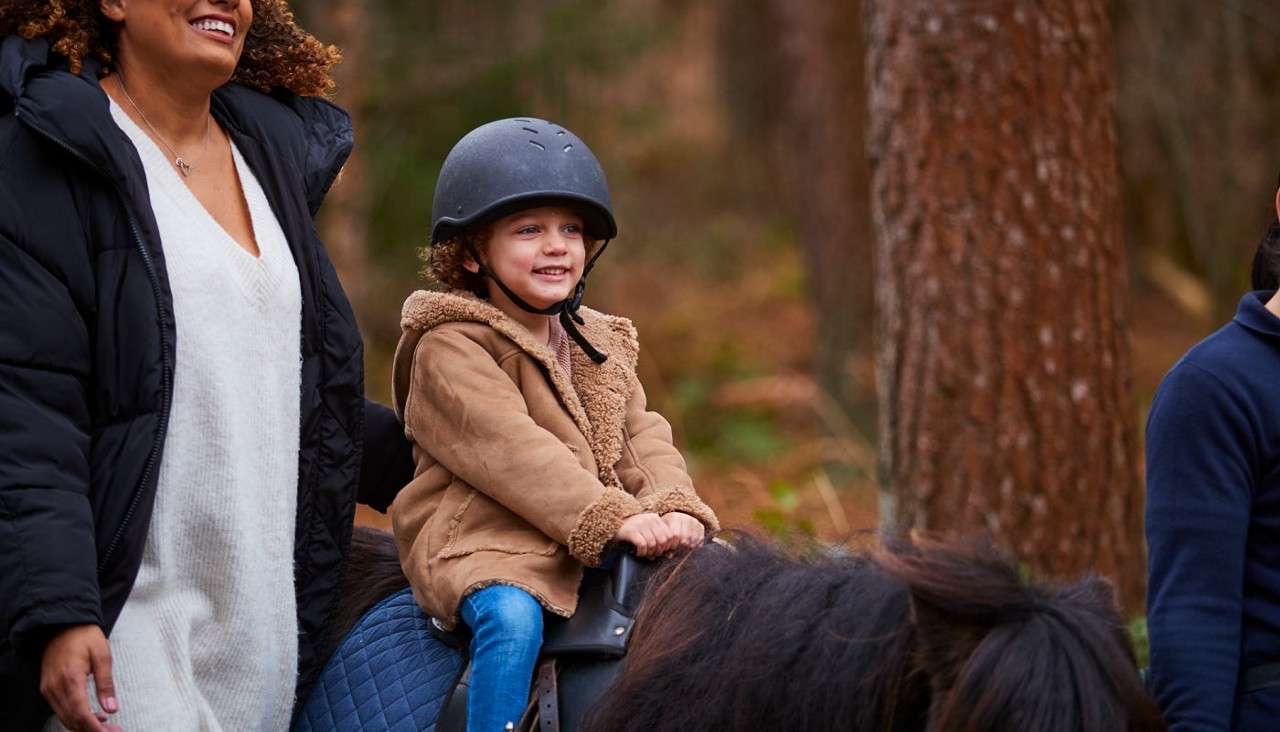 Child in a helmet rides a pony, smiling and gripping the saddle, while two adults walk beside, guiding along a wooded forest trail.