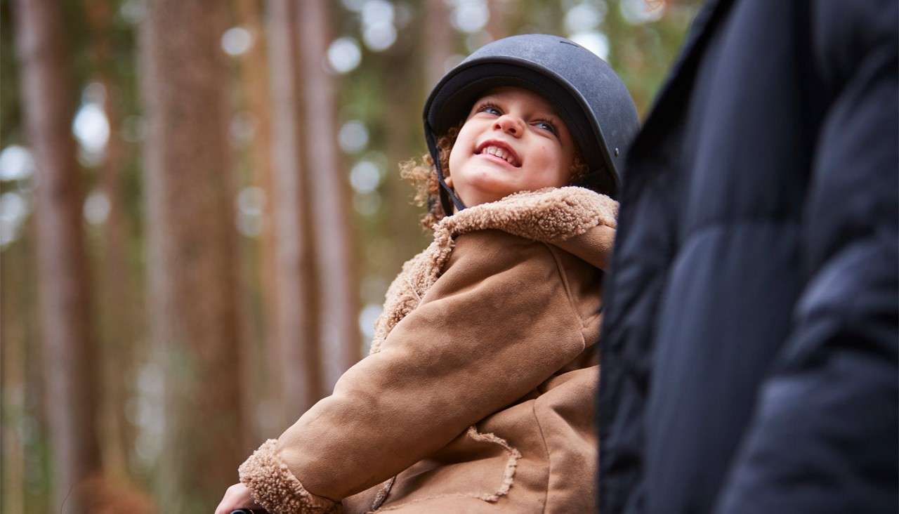 Child in a riding helmet and shearling coat smiles upward while sitting beside an adult; blurred tall trees fill the forest background.